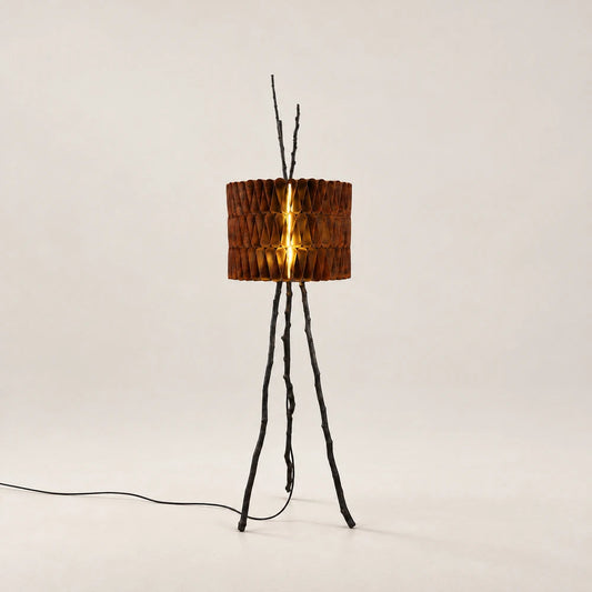Tudor Floor Lamp by Eva Szumilas, with three dark branch-like legs and a cylindrical shade of folded rust-toned metal illuminated from within.