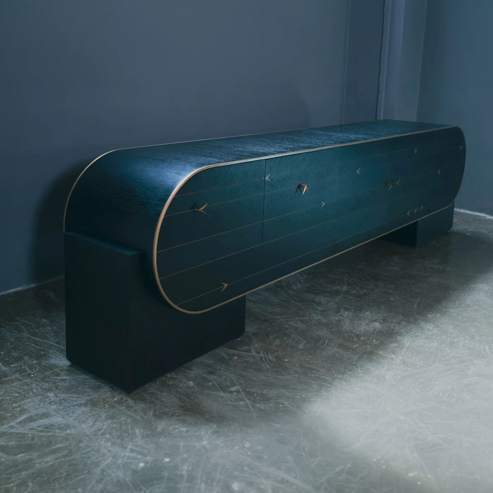 Angled view of the dark C01 Sideboard by Eva Szumilas, showing its curved case, gold-toned trim and geometric door details against a blue-grey wall.
