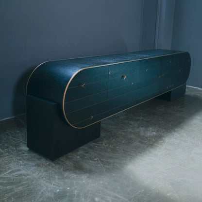 Angled view of the dark C01 Sideboard by Eva Szumilas, showing its curved case, gold-toned trim and geometric door details against a blue-grey wall.