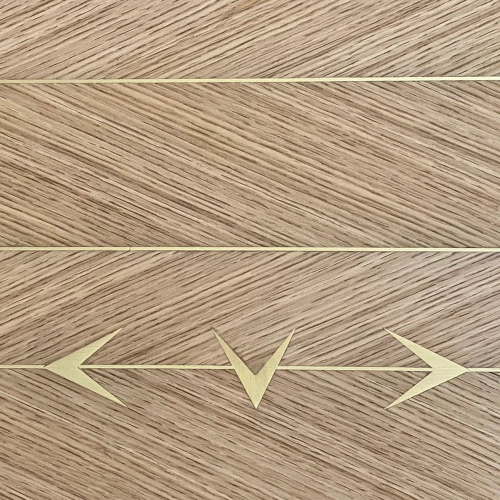 Macro detail of the oak DT01 Dining Table by Eva Szumilas, showing alternating diagonal wood grain, fine gold-toned lines and three directional arrow inlays.