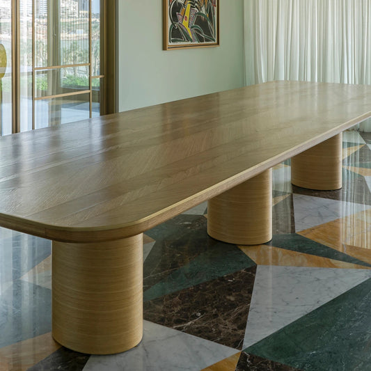 Oak DT01 Dining Table by Eva Szumilas in a bright interior, showing its long patterned wooden top and three cylindrical supports above a geometric marble floor.