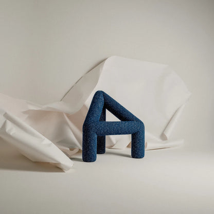 Blue A Chair by Garnier Pingree against folded white paper, with upholstered cylindrical elements forming an open triangular silhouette.