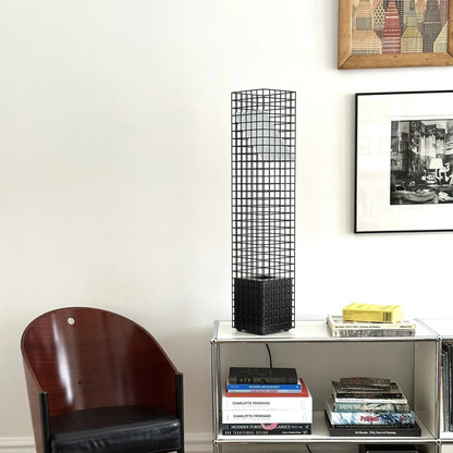 Unlit black Ami Table Lamp by Garnier Pingree on metal shelving, beside a wooden armchair and framed artwork.
