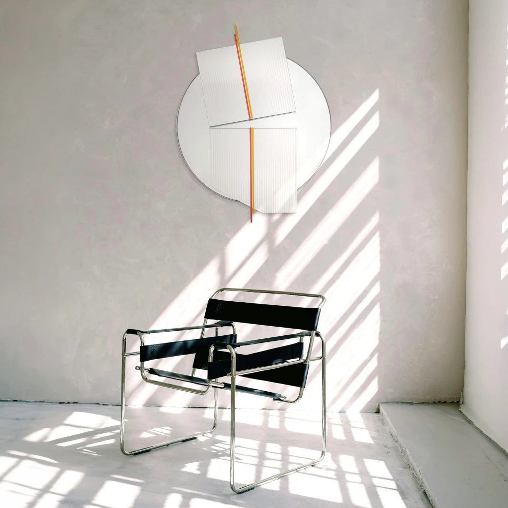 Large Claude Mirror by Garnier Pingree with red-orange and yellow rods, above a black tubular-frame armchair in an interior crossed by sunlight.