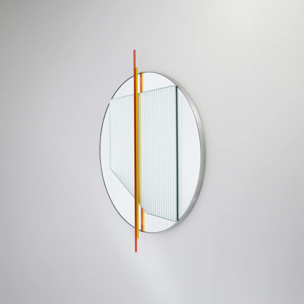 Angled view of the small Claude Mirror by Garnier Pingree, with red-orange and yellow rods crossing its circular mirror and ribbed glass panel.