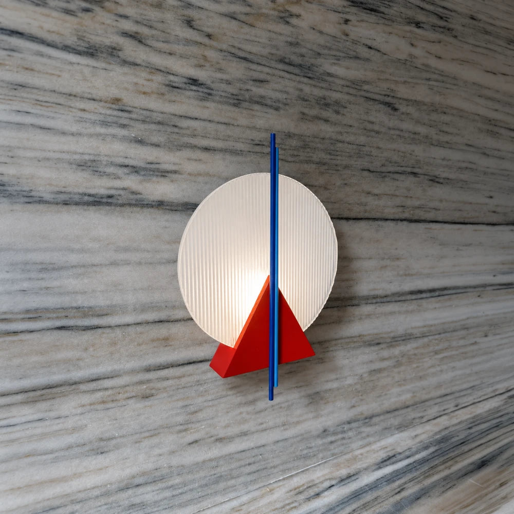 Angled view of the illuminated Claude Wall Lamp by Garnier Pingree, with a ribbed glass disc, red triangle and blue rods on veined stone.