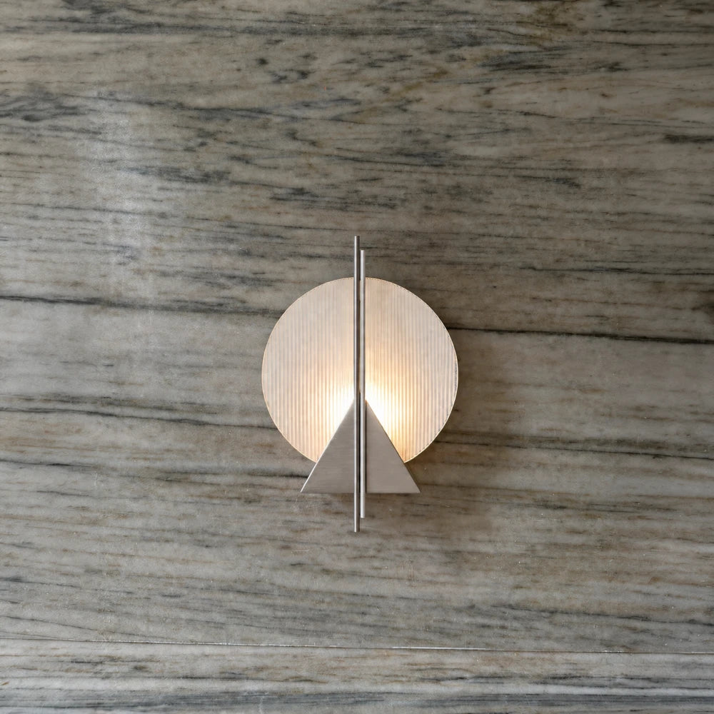 Claude Wall Lamp by Garnier Pingree on a veined stone wall, with a luminous ribbed glass disc, stainless steel triangle and vertical rods.