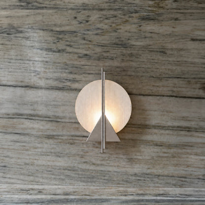 Claude Wall Lamp by Garnier Pingree on a veined stone wall, with a luminous ribbed glass disc, stainless steel triangle and vertical rods.