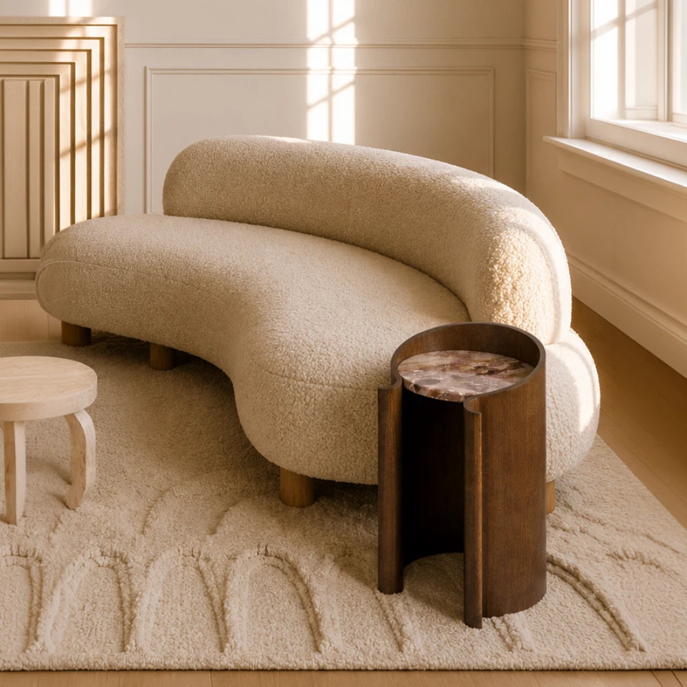 Dark wood Volute Side Table by Garnier Pingree beside a curved cream sofa, with a pink veined stone top in a sunlit interior.