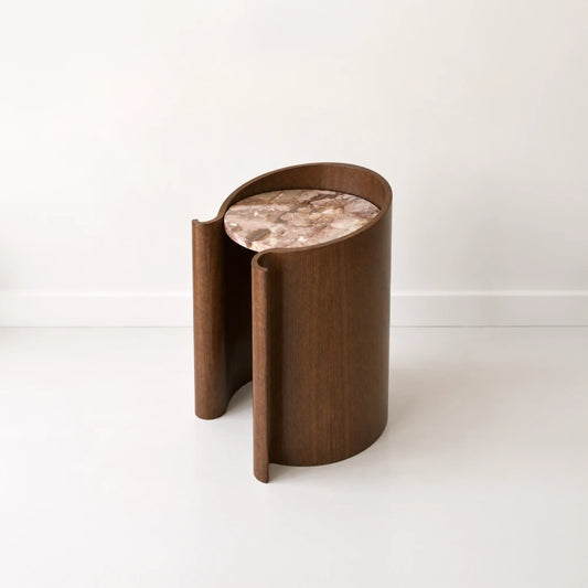 Three-quarter view of the Volute Side Table by Garnier Pingree, showing its dark wood grain, curled edges and circular pink veined stone top.