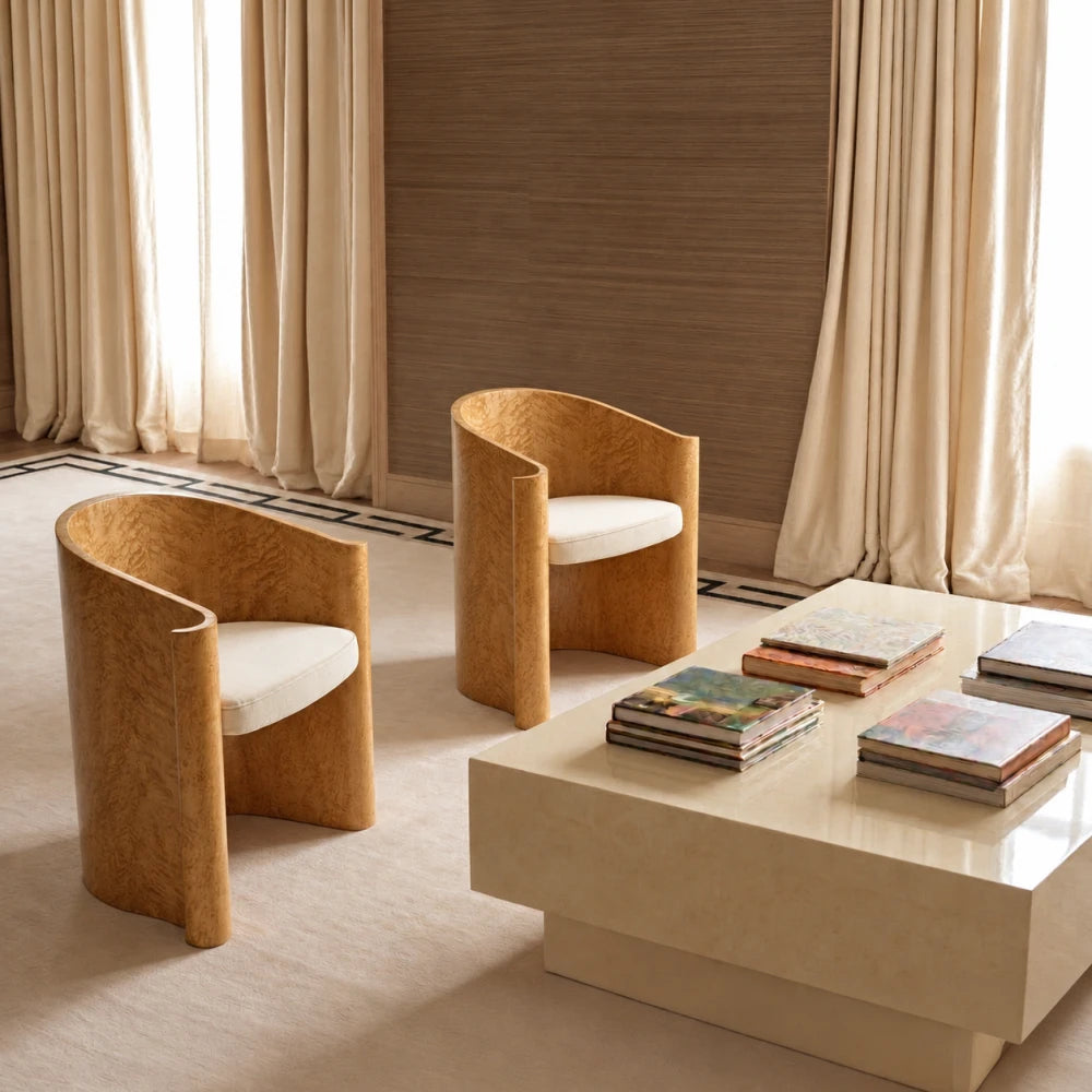 Two golden wood Volute Chairs by Garnier Pingree with cream seats, arranged beside a pale stone coffee table in a curtained interior.