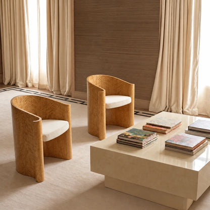 Two golden wood Volute Chairs by Garnier Pingree with cream seats, arranged beside a pale stone coffee table in a curtained interior.