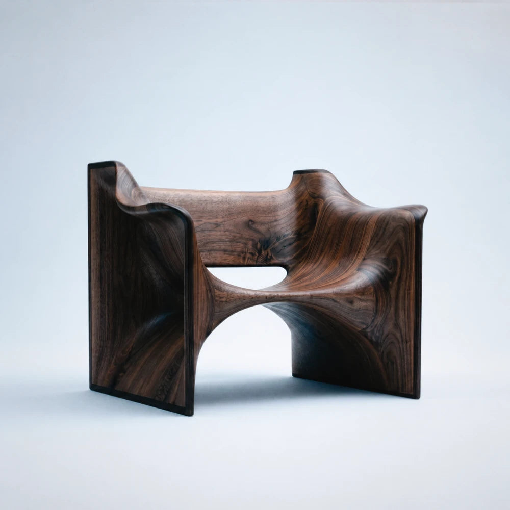 Three-quarter view of the Deus Walnut Chair by Studio Romell, revealing its flowing seat, architectural side panels and expressive walnut grain.