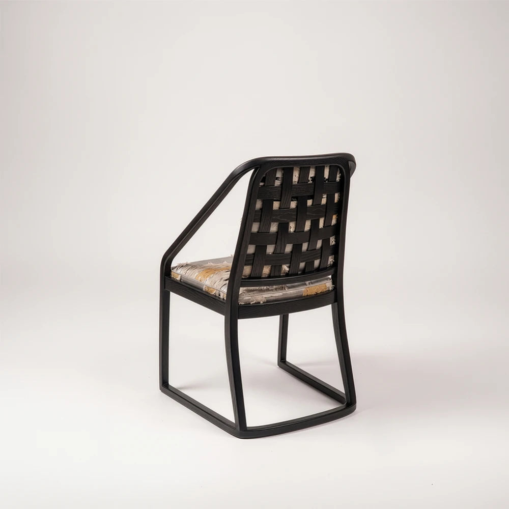 Rear three-quarter view of the Oblation Dining Chair by Studio Romell, showing its black wooden frame, interwoven backrest slats and patterned upholstered seat.
