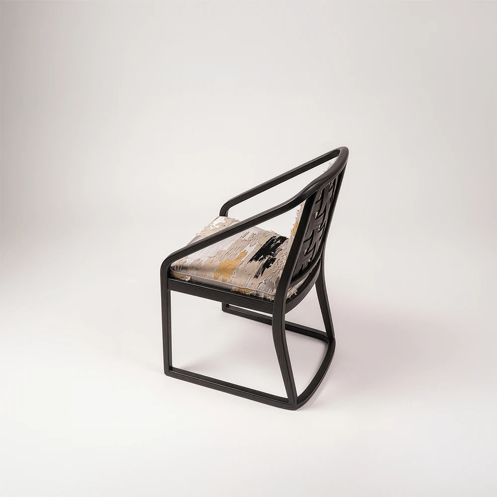 Angled side view of the Oblation Dining Chair by Studio Romell, showing its interwoven wooden backrest, sweeping armrests and grey upholstery with contrasting motifs.