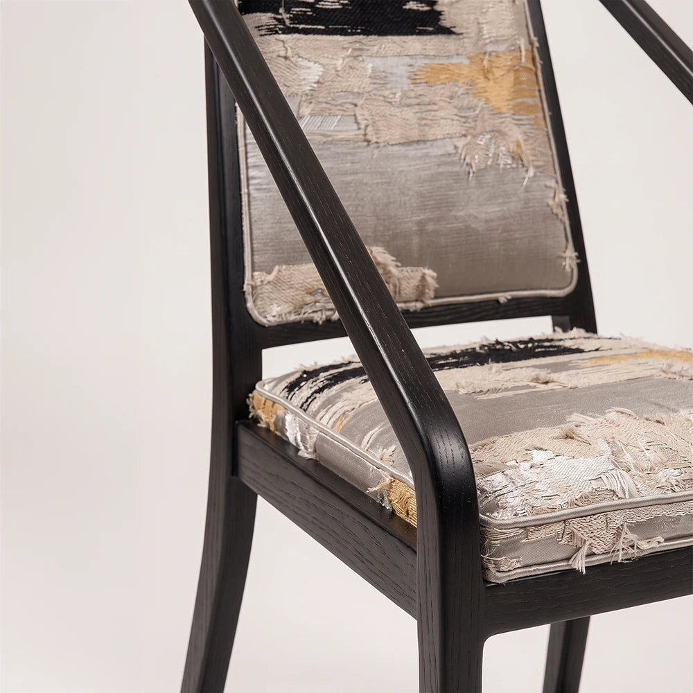 Detail of the Oblation Dining Chair by Studio Romell, showing the curved black wooden armrest, visible wood grain and piped edges of the textured cushions.